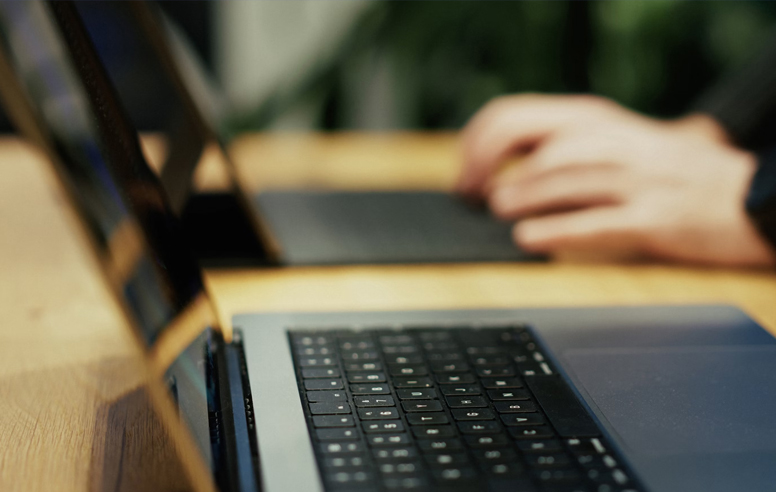 Close-up of hands typing on a laptop keyboard at a shared desk, with a second laptop partially visible in the foreground.