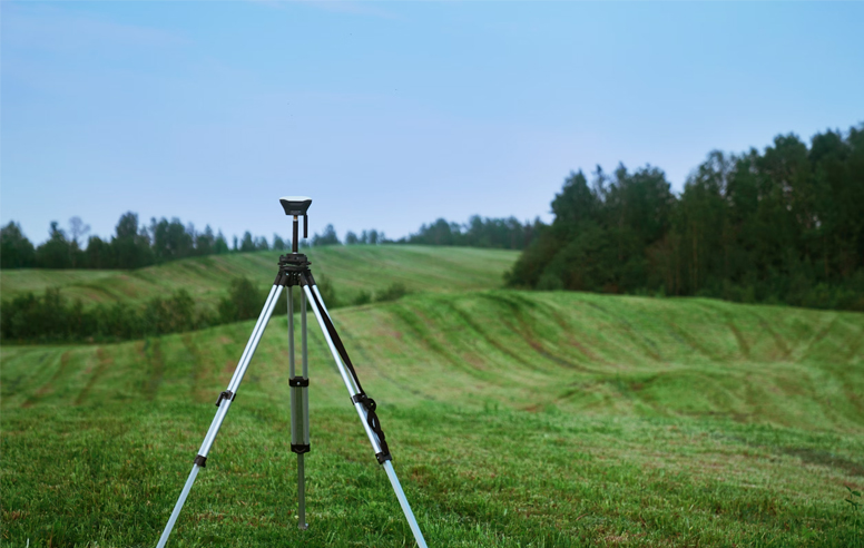 Surveying tripod standing in the middle of a wide, grassy field with tree-lined hills in the background under a clear blue sky.